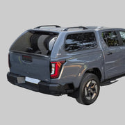 A dark gray pickup truck Travo with a hardtop canopy and black wheels, photographed from the rear three-quarter angle in an outdoor driveway in front of gray garage doors.